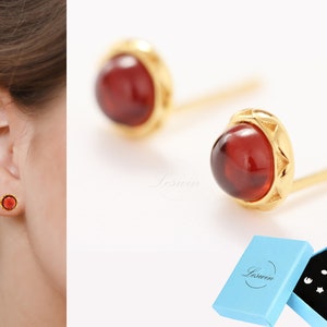May include: A pair of gold stud earrings with red gemstone accents. The earrings are shown in a close-up view, with one earring worn in a person's ear and the other earring displayed on a white surface. The earrings are packaged in a blue box with the brand name "Laswen" printed on the box.