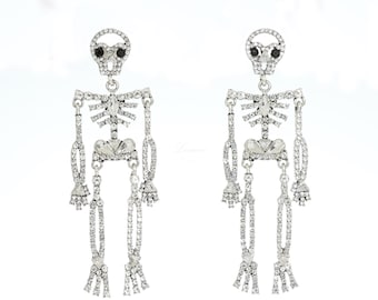 May include: A pair of silver-toned skeleton earrings. Each earring features a skull with black eyes and a body adorned with clear crystals. The earrings are designed to dangle, creating a unique and eye-catching accessory.