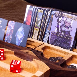 May include: A wooden card and dice organiser with two red dice and playing cards. The organiser has compartments for cards, dice, and tokens. The cards feature fantasy art. The dice are red with white spots.