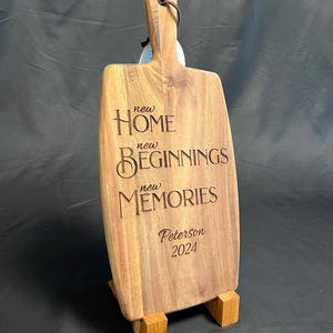 May include: A wooden chopping board with the text "new HOME new BEGINNINGS new MEMORIES Peterson 2024" engraved on it. The board is standing on a wooden stand.