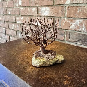 Op de afbeelding: Een boomsculptuur gemaakt van gedraaid koperdraad, met een dicht netwerk van takken en een brede, uitlopende basis van wortels. De draadboom is stevig verankerd op een enkele, lichtgekleurde, onregelmatig gevormde natuurstenen basis.