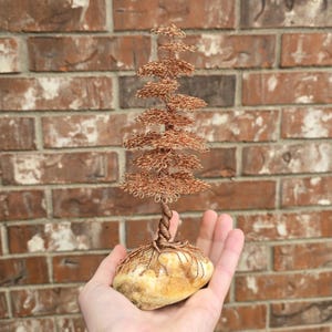 Puede incluir: Escultura artesanal de un árbol de alambre de cobre. El árbol presenta un dosel detallado y en capas y un tronco retorcido, enraizado en una base de piedra natural. El alambre de cobre tiene un cálido tono marrón rojizo.