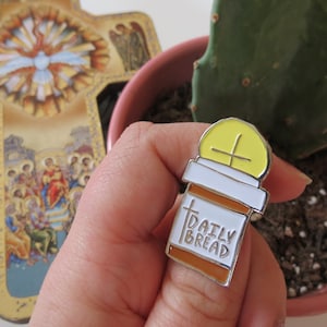 May include: A yellow and white enamel pin shaped like a pill bottle with a cross on top. The bottle says "Daily Bread" on the label.