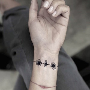 Puede incluir: Un diseño de tatuaje de margaritas negras en la muñeca. El tatuaje presenta tres flores de margarita conectadas por una fina línea negra. La muñeca es de piel clara y se ve una pulsera roja debajo.