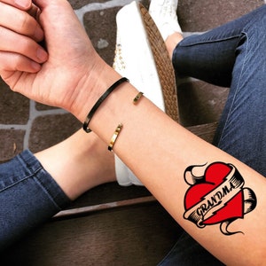May include: A red heart with a banner that says "Grandma" in black lettering. The heart is surrounded by black swirls. The tattoo is on a person's arm.