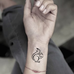 Könnte beinhalten: Ein schwarzes Tattoo im Linienstil eines Eichhörnchens auf einem Handgelenk. Das Eichhörnchen ist in einem stilisierten, minimalistischen Design dargestellt, mit einem gebogenen Schwanz und einem abgerundeten Körper. Das Tattoo befindet sich auf einem hellhäutigen Handgelenk.