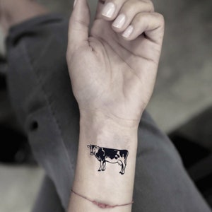 May include: A black and white cow temporary tattoo on a wrist. The cow design is a simple illustration with black spots and a white body. The tattoo is placed on the wrist of a person.