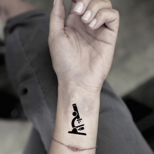 May include: A black microscope temporary tattoo is shown on a wrist. The tattoo is a simple, stylized illustration of a microscope, with a base, a tube, and a lens. The skin tone is light, and the background is blurred.