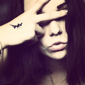 May include: A black silhouette tattoo of a fairy on a hand, with the fingers forming a peace sign over the face. The person has long dark hair and a silver bracelet.