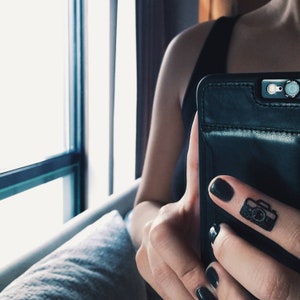 Könnte beinhalten: Schwarze Leder-Handyhülle mit Kameraobjektiv und Blitz. Auf dem Zeigefinger befindet sich ein schwarzes Kamera-Tattoo. Die Person trägt ein schwarzes Tanktop. Der Hintergrund umfasst ein Fenster und ein helles Sofa.
