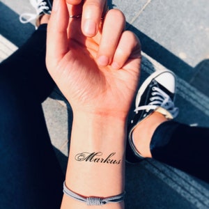 May include: A wrist tattoo with the name "Markus" in elegant cursive script. The tattoo is on a person's wrist, with a gray bracelet below it. Black and white sneakers and dark pants are visible in the background.