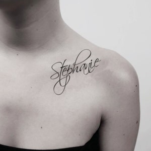 May include: A close-up of a person's shoulder with a cursive tattoo of the name "Stephanie" in black ink.