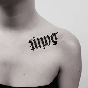 May include: A black tattoo on the upper chest and shoulder area. The tattoo features the word "saint" in a gothic font. The skin tone is light, and the background is plain.