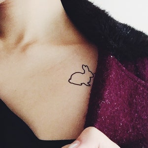 May include: A black outline of a rabbit tattoo on the skin. The person is wearing a black top and a dark purple coat with black fur trim.