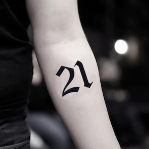 Könnte beinhalten: Schwarzes temporäres Tattoo der Zahl 21 in einem gotischen Schriftstil. Das Tattoo wird auf einem Arm mit hellem Hautton dargestellt. Der Hintergrund ist verschwommen mit hellem Licht.