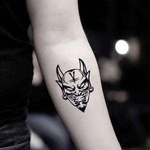 Peut inclure: Tatouage temporaire noir et blanc d'un visage de diable. Le motif représente un diable cornu avec un sourire sinistre, une marque en forme de V sur le front et une barbe triangulaire. Le tatouage est appliqué sur le bras.