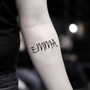 May include: A black temporary tattoo on an arm. The tattoo features the name "EMMA" written in a handwritten style. The arm is fair-skinned, and the background is blurred.