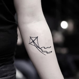 May include: A black and white line drawing of a kite flying in the air. The kite has a simple design with a tail.