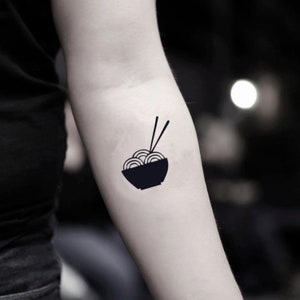 Può includere: Tatuaggio temporaneo nero di una ciotola di noodles con bacchette. La ciotola è piena di noodles circolari e due bacchette sono posizionate sopra. Il design è semplice e minimalista, perfetto per gli amanti del cibo.