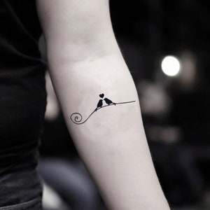May include: A black tattoo design featuring two birds perched on a line, with a heart between them. The line curves into a spiral. The tattoo is on a person's arm.
