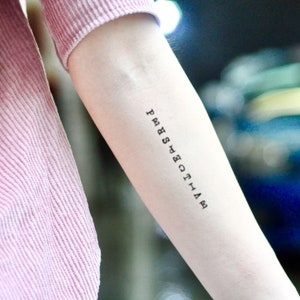 Puede incluir: Primer plano de un brazo con un pequeño tatuaje negro. El tatuaje es una línea vertical de texto que dice "PERSPECTIVE" en una fuente simple, sin serifa. El brazo es pálido y la persona lleva una camisa rosa de manga larga acanalada.