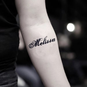 May include: A black tattoo on an arm displays the name "Melissa" in elegant cursive script. The word is in a flowing font with decorative swirls, set against a blurred background.