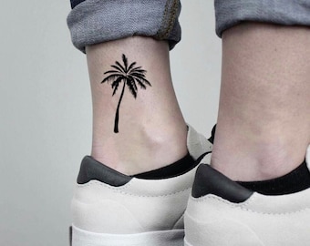 May include: A black palm tree temporary tattoo is visible on an ankle. The tattoo is a simple silhouette of a palm tree. The person is wearing white trainers with black accents and blue jeans.