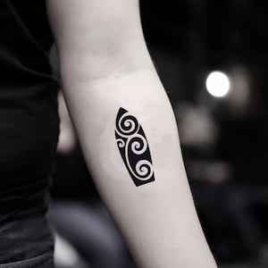 Puede incluir: Un tatuaje temporal negro con forma de tabla de surf. El diseño de la tabla de surf presenta patrones de olas estilizadas y arremolinadas. El tatuaje está aplicado en el brazo, con un fondo borroso.