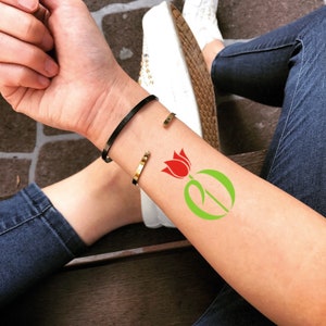 May include: A black and gold cuff bracelet set. The black bracelet is on top, and the gold bracelet is below. A red tulip tattoo with green leaves is visible on the arm. The person is wearing blue jeans and white sneakers.