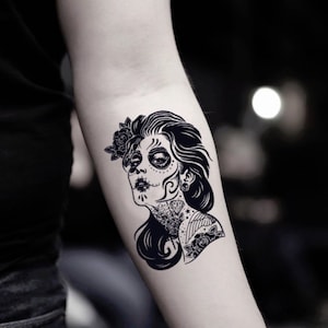 May include: Black and white temporary tattoo of a woman's face, styled in the Day of the Dead aesthetic. The design features intricate makeup, a flower in the hair, and detailed neck and shoulder tattoos.
