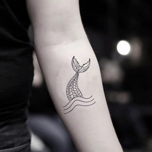 Könnte beinhalten: Ein schwarz-weißes temporäres Tattoo eines Meerjungfrauenschwanzes. Der Schwanz ist mit komplizierten Mustern versehen und erhebt sich aus zwei wellenförmigen Linien, die Wasser darstellen. Das Design befindet sich auf dem Arm.