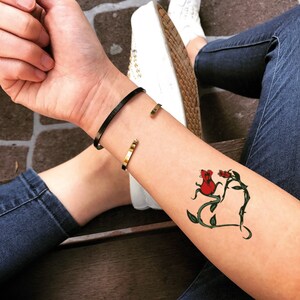 May include: A black cuff bracelet and a gold cuff bracelet are worn on the wrist. A temporary tattoo of two red roses with green vines and leaves is visible on the forearm.