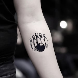 May include: A black and white temporary tattoo of a bowling ball and pins. The bowling ball is centered in front of five bowling pins. The pins are arranged in a triangular formation. The tattoo is on a person's arm.