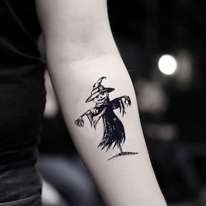 Peut inclure: Tatouage temporaire noir et blanc d'un épouvantail portant un chapeau avec un visage souriant. L'épouvantail a les bras tendus et se tient sur un petit bout de terre. Le tatouage est appliqué sur le bras.