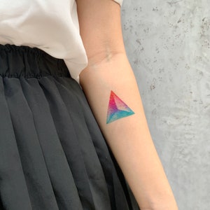 May include: A colorful geometric tattoo in the shape of a triangle, with a gradient of red, blue, and purple hues. The tattoo is on the forearm of a person wearing a white top and black skirt.
