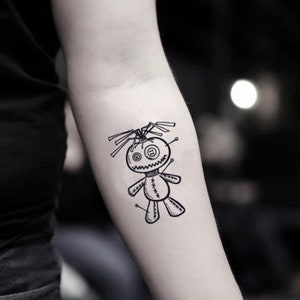 May include: A black and white tattoo of a voodoo doll with button eyes, a stitched mouth, and pins. The doll has a stitched body and limbs, with hair-like lines extending from its head. The tattoo is on a person's arm.
