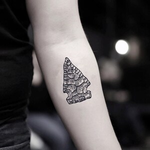 May include: A black and white temporary tattoo of an arrowhead. The arrowhead design features detailed shading and a textured appearance, resembling a stone tool. The tattoo is applied to the arm.
