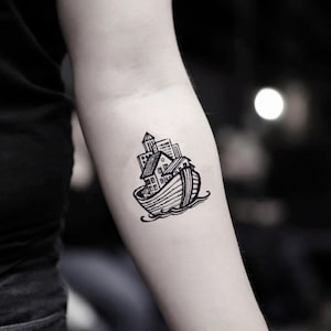 May include: A black and white tattoo of a boat carrying buildings, houses, and a tower. The boat is on water, and the design is a line drawing. The tattoo is on a person's arm.