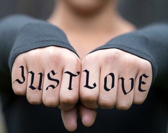May include: Close-up of two hands with the knuckles displaying the words "JUST LOVE" in a gothic font. The hands are positioned in front of a blurred background, with the person wearing a black long-sleeved shirt.