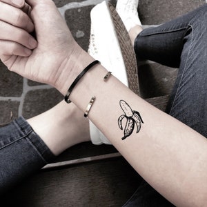 Könnte beinhalten: Ein temporäres Tattoo in Schwarz-Weiß einer geschälten Banane ist auf einem Arm zu sehen. Zwei offene Armreifen, einer schwarz und einer goldfarben, werden am Handgelenk getragen. Die Person trägt eine blaue Jeans und weiße Turnschuhe.