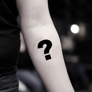 Könnte beinhalten: Ein schwarzes Fragezeichen-Tattoo auf einem Arm. Das Tattoo ist ein einfaches, kühnes Design mit einem gebogenen oberen Teil und einem quadratischen Punkt darunter. Der Hintergrund ist verschwommen, mit einem weichen, unscharfen Licht.