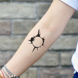 May include: A black temporary tattoo featuring a circular design with spiky extensions, reminiscent of a sun or burst. The tattoo is on the forearm, offering a minimalist and bold look. The design is simple but striking.