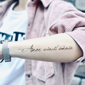 May include: A temporary tattoo on an arm displays the phrase "Amor vincit omnia" in elegant cursive script. The arm is adorned with a silver watch with a mesh band. The person is wearing a pink corduroy shirt and a white t-shirt.