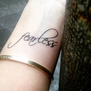 May include: A black script tattoo on the wrist reads "fearless." A gold-colored bracelet is worn below the tattoo. The image is a close-up shot, with a blurred background.