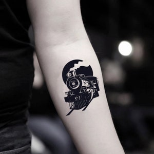 May include: A black and white temporary tattoo of a vintage steam train. The train is set against a dark circular background, with smoke billowing from the smokestack. The tattoo is applied to the arm.