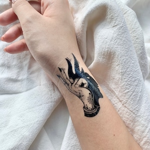 Peut inclure: Tatouage temporaire noir représentant une main tenant une silhouette de loup. Le motif comprend un pentagramme, une toile d'araignée et d'autres détails. Le tatouage est appliqué sur le poignet.