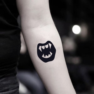 May include: A black temporary tattoo depicting a stylized mouth with fangs. The tattoo is on the forearm, set against a dark background. The design is a striking, graphic representation of vampire teeth.