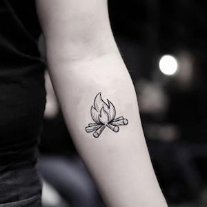 Könnte beinhalten: Schwarz-Weiß-Illustration eines Lagerfeuer-Tattoos. Das Design zeigt stilisierte Flammen über gekreuzten Baumstämmen, wodurch ein einfaches, aber detailliertes Bild entsteht. Das Tattoo befindet sich auf dem Arm einer Person.