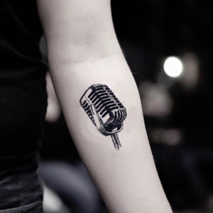 Könnte beinhalten: Schwarz-weiß Tattoo eines Vintage-Mikrofons auf einem Arm. Das Mikrofon ist detailliert mit parallelen Linien und einer abgerundeten Oberseite, vor einem verschwommenen Hintergrund.