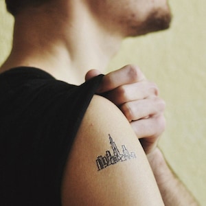 May include: A black and white tattoo of a city skyline on a person's shoulder. The tattoo features a detailed outline of buildings and skyscrapers, with a prominent tower in the center. The person is wearing a black shirt.